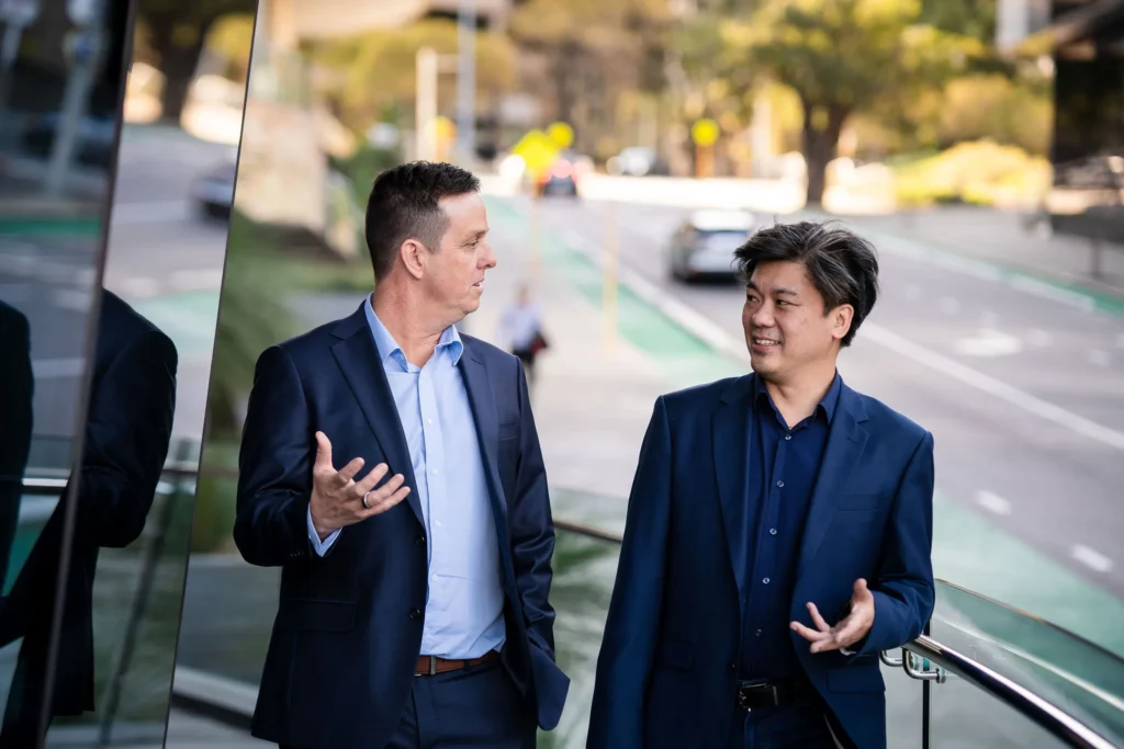 anaging Directors in professional attire walking along a sunny city sidewalk, engaged in an active, collaborative discussion.