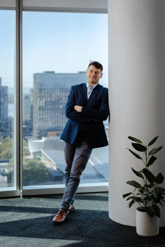 A professional standing confidently by a floor-to-ceiling window in a modern high-rise office, with a sweeping city skyline in the background.